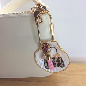 May include: A white handbag charm featuring a seashell base with gold-painted edges. The shell displays a painted handbag graphic with a leopard print pattern, a small pink bow, a pink tassel, a white pearl bead, and a red patterned bead.