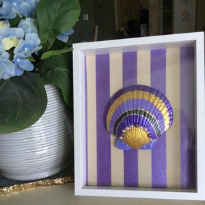 May include: A white shadow box frame containing a seashell painted in purple, gold, and black stripes. The shell is decorated with a row of small white and dark purple beads along its center. The background features vertical purple and cream stripes.