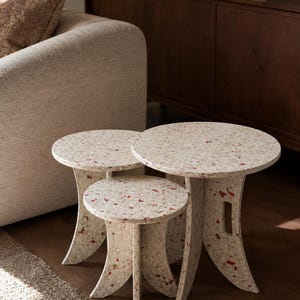 Puede incluir: Un juego de tres mesas nido con tableros circulares y patas curvas, fabricadas con un material compuesto de color blanco roto moteado con incrustaciones de motas rojas y marrones en toda la superficie y los componentes.