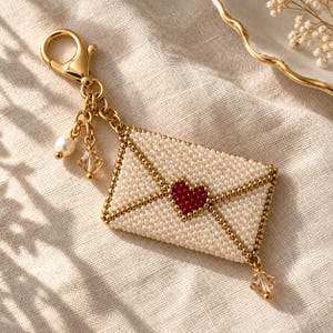 May include: A gold-toned metal keychain featuring a beaded envelope charm. The envelope is crafted from small white beads with a central red heart design. It includes a gold lobster clasp, a single white pearl bead, and two faceted champagne-colored crystal charms.