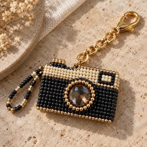 May include: A camera-shaped keychain crafted from black, cream, and gold-toned beads. The center features a large, faceted dark crystal lens. It includes a gold-toned metal chain with a lobster clasp and a small beaded loop attached to the side.
