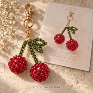 May include: A beaded cherry bag charm featuring two round red crystal spheres attached to green crystal stems and leaves, connected to a gold-tone metal lobster clasp. Beside it lies a card labelled Sunday Bloom Atelier Cerise The Crystal Cherry Bag Charm.