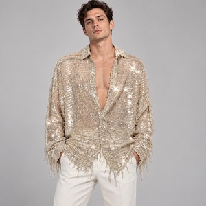 May include: A long-sleeved button-down shirt covered in shimmering gold sequins and delicate fringe detailing. The garment features a classic collar, a buttoned front placket, and a loose fit, paired with plain white trousers.