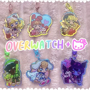 May include: Six acrylic keychains featuring colorful anime-style characters and Sanrio mascots, each with a matching plastic clasp. The text OVERWATCH + is printed in a rainbow gradient font, accompanied by a pink cat icon with a bow.