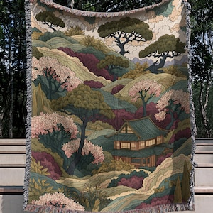 May include: A woven throw blanket featuring a detailed landscape illustration of a traditional house nestled among rolling hills, pine trees, and blooming cherry blossom trees in shades of green, pink, burgundy, and cream with a fringed edge.