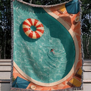 Peut inclure: Un plaid tissé représentant une vue plongeante sur une piscine à l'eau turquoise, une bouée rayée rouge et blanche, une personne qui nage, et une terrasse en carrelage orange avec des parasols bleus et des chaises longues jaunes.