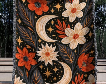 Könnte beinhalten: Eine gewebte Wohndecke mit schwarzem Hintergrund und einem wiederkehrenden Muster aus orangefarbenen und cremefarbenen Blumen, dunkelgrauen Blättern, Mondsicheln und vierzackigen Sternen. Die Decke hat einen Fransenrand oben und unten.