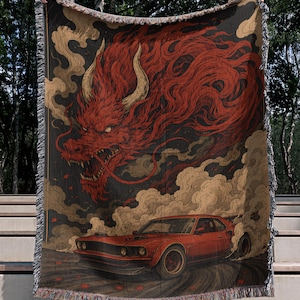 May include: A woven throw blanket featuring a graphic illustration of a red muscle car drifting on a road, surrounded by clouds of smoke. Above the car, a large, detailed red dragon with horns emerges from the smoke. The blanket has fringed edges.
