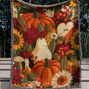 Op de afbeelding: Een geweven plaid met een levendig herfstpatroon van oranje pompoenen, rode appels, een witte peer, gele zonnebloemen, rode bessen, paddenstoelen en kleurrijke herfstbladeren, afgewerkt met franjes aan de boven- en onderkant van de deken.