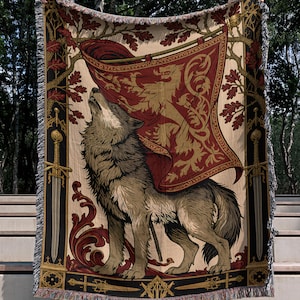 Peut inclure: Un plaid tissé présentant une illustration centrale d'un loup gris hurlant vers une bannière rouge et dorée. Le motif comprend des épées verticales sur les côtés, des feuilles de chêne et des volutes ornées dans des tons rouge, or et noir.