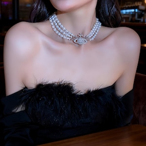 May include: A three-strand pearl choker necklace featuring a central silver-toned orb pendant encrusted with clear crystals. The necklace is worn around the neck, showcasing the layered white pearls and the detailed metallic orb centerpiece.