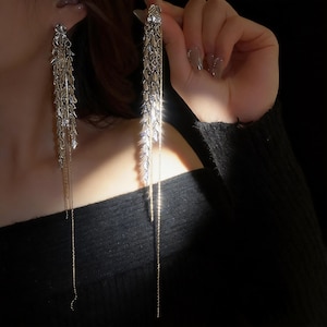 May include: A pair of long, silver-toned dangle earrings featuring a cascading wheat-like design composed of multiple metallic leaf-shaped elements. Each earring includes a delicate, thin chain that hangs vertically from the main structure.