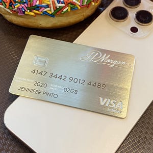 Op de afbeelding: Een zilverkleurige metalen J.P. Morgan Visa Infinite creditcard met de naam Jennifer Pinto, kaartnummer 4147 3442 9012 4489 en verloopdatum 02/28, liggend op een witte smartphone naast een donut met kleurrijke hagelslag.