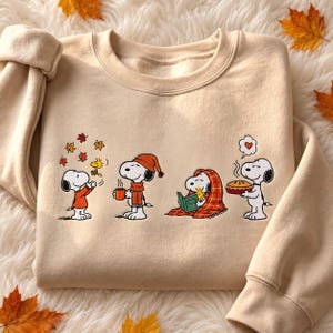 May include: A beige crewneck sweatshirt featuring a graphic print of Snoopy and Woodstock in four autumn-themed scenes: playing with falling leaves, holding a mug, reading under a plaid blanket, and holding a pie with a heart speech bubble.