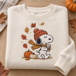 Peut inclure: Un sweat-shirt à col rond couleur crème avec une broderie représentant un chien blanc portant un bonnet en tricot orange et une écharpe jaune moutarde. Le chien tient une tasse de café, entouré de feuilles d'automne et de deux citrouilles.