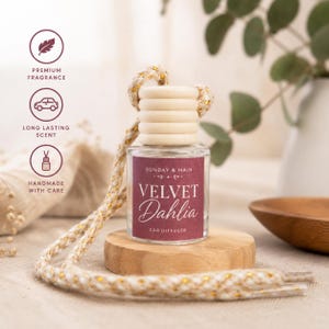 May include: A clear glass car diffuser bottle with a light wood cap and a braided cord. The label is dark red with white text reading SUNDAY & MAIN, VELVET Dahlia, and CAR DIFFUSER. Three circular icons on the left show a leaf, a car, and a bottle.
