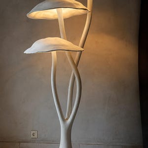 May include: A floor lamp shaped like three mushrooms with a white, textured finish. The lamp features a wide, circular base and three stems that branch upward, each topped with a wide, curved cap that emits a warm, soft glow from underneath.