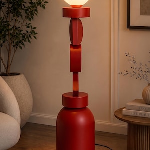 May include: A floor lamp featuring a matte red cylindrical base and a vertical stack of geometric red shapes. The lamp is topped with a spherical white glass globe light bulb that emits a warm glow. A black power cord extends from the base.