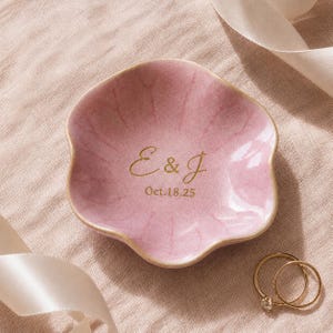 Puede incluir: Un plato decorativo de cerámica rosa con forma de flor y un acabado de esmalte brillante con efecto craquelado. El centro presenta un texto dorado que dice E & J y Oct. 18.25. El plato tiene un borde festoneado y una superficie lisa.