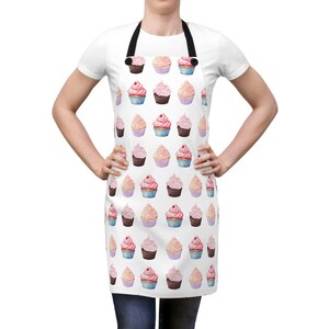 May include: A white apron featuring a repeating pattern of watercolor-style cupcakes in shades of pink, brown, and blue. The apron has black neck and waist straps and a straight-cut top edge, displaying a variety of frosted cupcake illustrations.
