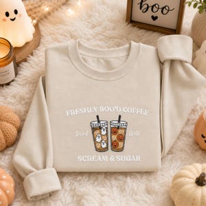 May include: A beige crewneck sweatshirt featuring an embroidered graphic of two iced coffee cups. One cup contains white ghost shapes, the other orange pumpkin faces. Text reads: FRESHLY BOO'D COFFEE, Served With, SCREAM & SUGAR.