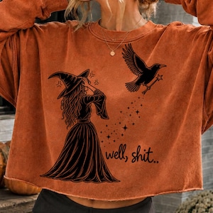 Pode incluir: Uma camisola de manga comprida em laranja queimado com uma impressão gráfica preta de uma bruxa com chapéu pontiagudo e um corvo a voar que segura uma varinha mágica. O design inclui um rasto de estrelas e o texto well, shit.. em preto.