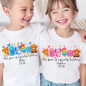 May include: Two white t-shirts featuring a colorful graphic of a pizza slice, jellyfish, axolotl, popsicle, ice cube, and highland cow. The text reads the year of squishy hunting with the names Chloe or Sophia and the year 2026 printed below.