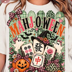 May include: A white t-shirt featuring a graphic with the text MAHJ-O-WEEN in orange and pink. The design includes three mahjong tiles, a jack-o'-lantern, a leopard print pumpkin, two ghosts with pink bows, a witch hat, and plaid bows.