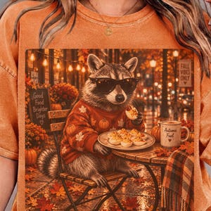 Puede incluir: Una camiseta naranja de cuello redondo con el gráfico de un mapache con gafas de sol y un suéter de calabazas, sentado a una mesa comiendo huevos rellenos. La imagen incluye una taza con la etiqueta Autumn Fuel y un cartel que dice Good Food Brighter Days.