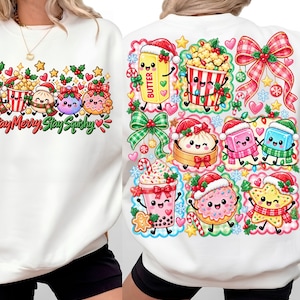 Puede incluir: Una sudadera blanca con un estampado gráfico colorido de temática navideña. El diseño incluye personajes de dibujos animados como mantequilla, palomitas, dumplings y una bebida, todos con gorros de Papá Noel, lazos, corazones y el texto Stay Merry Stay Squishy.