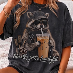 May include: A charcoal grey t-shirt featuring a graphic of a raccoon wearing a baseball cap and sunglasses, holding an iced coffee with a straw. The raccoon wears an apron with hair styling tools and the text "literally just a hairstylist".