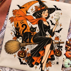 May include: A beige t-shirt featuring a graphic illustration of a witch in a black dress and pointed hat sitting on a broomstick. A black cat, a crescent moon, bats, a haunted house, and two jack-o'-lanterns are depicted in orange and black.