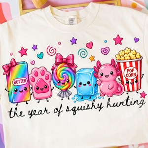 May include: A cream-coloured t-shirt featuring a colourful graphic of cartoon characters including a rainbow butter stick, a pink paw, a rainbow lollipop, a blue ice cube, a pink axolotl, and popcorn with the text the year of squishy hunting.