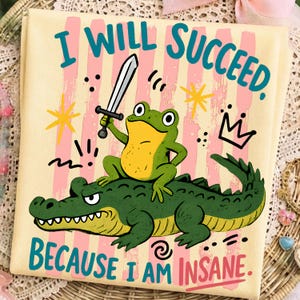 Puede incluir: Una camiseta de color amarillo claro con una rana verde que sostiene una espada mientras monta un caimán verde. El texto "I WILL SUCCEED. BECAUSE I AM INSANE." está impreso en azul verdoso y rosa. El fondo tiene rayas rosas verticales y estrellas amarillas.