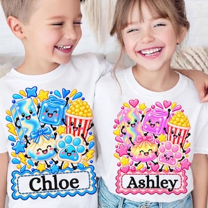 May include: Two white t-shirts featuring colourful cartoon graphics of a rectangular block, a cube, a popcorn bucket, a drawstring pouch, and a paw print. The left shirt includes the name Chloe, and the right shirt includes the name Ashley.