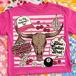 Może przedstawiać: Jaskraworóżowy t-shirt z centralną grafiką czaszki krowy w panterkę. Wzór zawiera czapkę z daszkiem z napisem "On My Husband's Top", symbol pika z napisem "Livin' On That Blue Collar Dollar" oraz metkę z napisem "Blue Collar Brat".