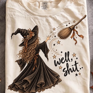 May include: A cream-coloured t-shirt featuring a graphic of a witch in a black dress and hat with flowers, reaching toward a flying broomstick. The design includes small stars and the text "well, shit.." in black script. The shirt has a Comfort Colors label.