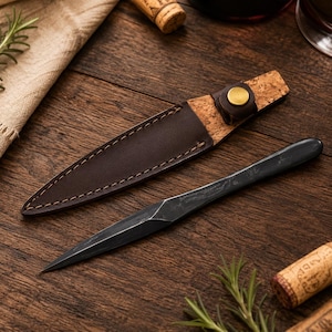 Puede incluir: Una herramienta puntiaguda de metal gris oscuro con una hoja facetada y una funda a juego. La funda está fabricada con una combinación de cuero marrón y corcho, con un cierre de botón a presión de metal color latón en el extremo superior.