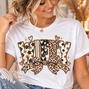 Peut inclure: Un t-shirt blanc à manches courtes avec le mot WIFEY écrit en grandes lettres majuscules. Chaque lettre présente un motif unique, notamment floral, rayé, à cœurs et léopard. Un grand nœud léopard est placé sous le texte avec des petits cœurs.