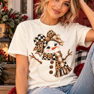 Peut inclure: Un t-shirt à manches courtes couleur crème avec un motif de bonhomme de neige portant un grand nœud et une écharpe à motifs léopard et à carreaux. Le bonhomme de neige a un nez en carotte, trois boutons marron et des bras en bâton.
