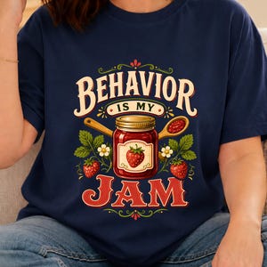Può includere: Una t-shirt blu navy con una stampa grafica di un barattolo di vetro di marmellata di fragole, due cucchiai di legno e piante di fragole. Il testo recita BEHAVIOR IS MY JAM in caratteri decorativi con accenti di colore rosso, bianco e verde.