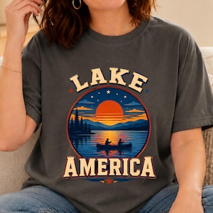 Peut inclure: Un t-shirt gris anthracite à manches courtes avec un motif circulaire représentant un coucher de soleil sur un lac, un canoë et deux personnes pagayant. Le texte LAKE est imprimé en arc de cercle crème au-dessus, et AMERICA en dessous.