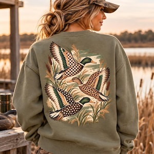 Peut inclure: Un sweat-shirt à col rond vert olive avec un motif au dos représentant trois canards en vol. Chaque canard présente un motif distinct sur ses ailes, incluant des carreaux, des taches de léopard et des pois, entouré de roseaux.