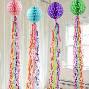 May include: Four hanging honeycomb paper spheres in pink, light blue, purple, and green. Each sphere features a long, cascading tail of multi-coloured paper ribbons in shades of red, orange, yellow, green, blue, and purple, creating a jellyfish effect.