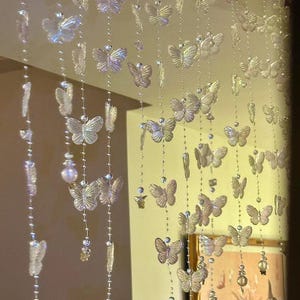以下が含まれることがあります： 透明で虹色に輝く蝶の形をしたビーズが連なる、複数の垂直なストランドで構成された吊り下げ式のルームディバイダーカーテン。蝶は細い金属製のチェーンに沿って繰り返しパターンで配置されており、きらめく半透明の効果を生み出しています。