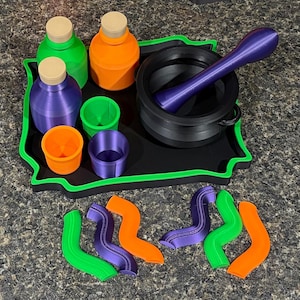 Pode incluir: Uma bandeja preta com borda verde cont&eacute;m tr&ecirc;s garrafas com rolhas bege em verde, laranja e roxo, tr&ecirc;s copos pequenos em verde, laranja e roxo, e um almofariz preto com pil&atilde;o. Seis formas onduladas em verde, laranja e roxo est&atilde;o dispostas abaixo.
