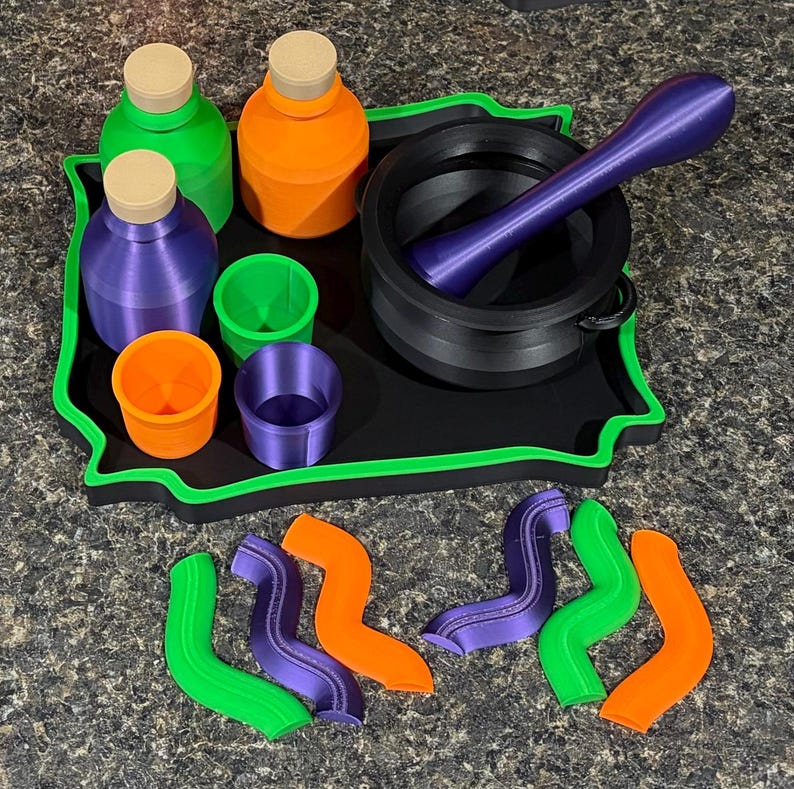 Pode incluir: Uma bandeja preta com borda verde cont&eacute;m tr&ecirc;s garrafas com rolhas bege em verde, laranja e roxo, tr&ecirc;s copos pequenos em verde, laranja e roxo, e um almofariz preto com pil&atilde;o. Seis formas onduladas em verde, laranja e roxo est&atilde;o dispostas abaixo.