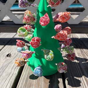 May include: A bright green, cone-shaped plastic tree stand with multiple holes, holding numerous Dum Dums lollipops in various colorful wrappers. The lollipops are inserted into the tree, creating a display of assorted candy flavors.