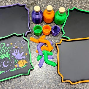 Pode incluir: Um conjunto de quatro tabuleiros retangulares com bordas recortadas em roxo, verde e laranja, tr&ecirc;s garrafas pequenas com rolhas, tr&ecirc;s copos pequenos e v&aacute;rias pe&ccedil;as de pl&aacute;stico curvas em roxo, laranja e verde, al&eacute;m de um tabuleiro com um gr&aacute;fico de Halloween.
