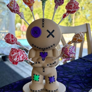 Pode incluir: Uma figura de boneco vudu texturizada em tom bronzeado com um olho de botão roxo, um olho em forma de X preto e uma boca costurada. O boneco apresenta um remendo de coração roxo no peito, remendos verdes e roxos nos joelhos e pirulitos.